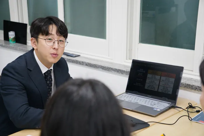 정장 차림의 담당자가 책상 맞은편 학생과 마주 앉아 노트북 화면의 표를 가리키며 설명하는 모습