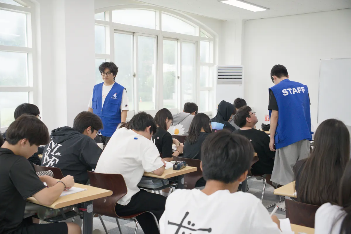강의실에 앉은 학생들이 시험지를 푸는 동안 조끼를 입은 담당자들이 감독하는 모습