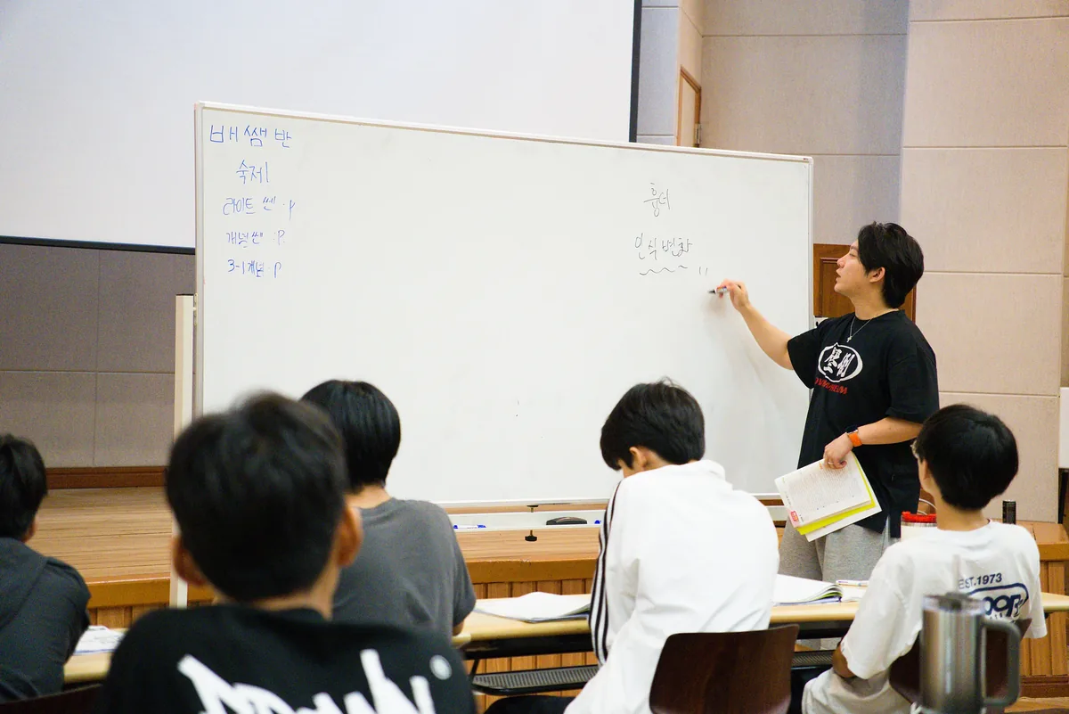 강사가 화이트보드에 판서하며 수업하고 학생들이 자리에 앉아 듣는 모습